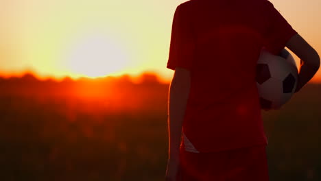 Young-boy-football-player-in-a-red-t-shirt-is-with-a-soccer-ball-on-the-field-at-sunset
